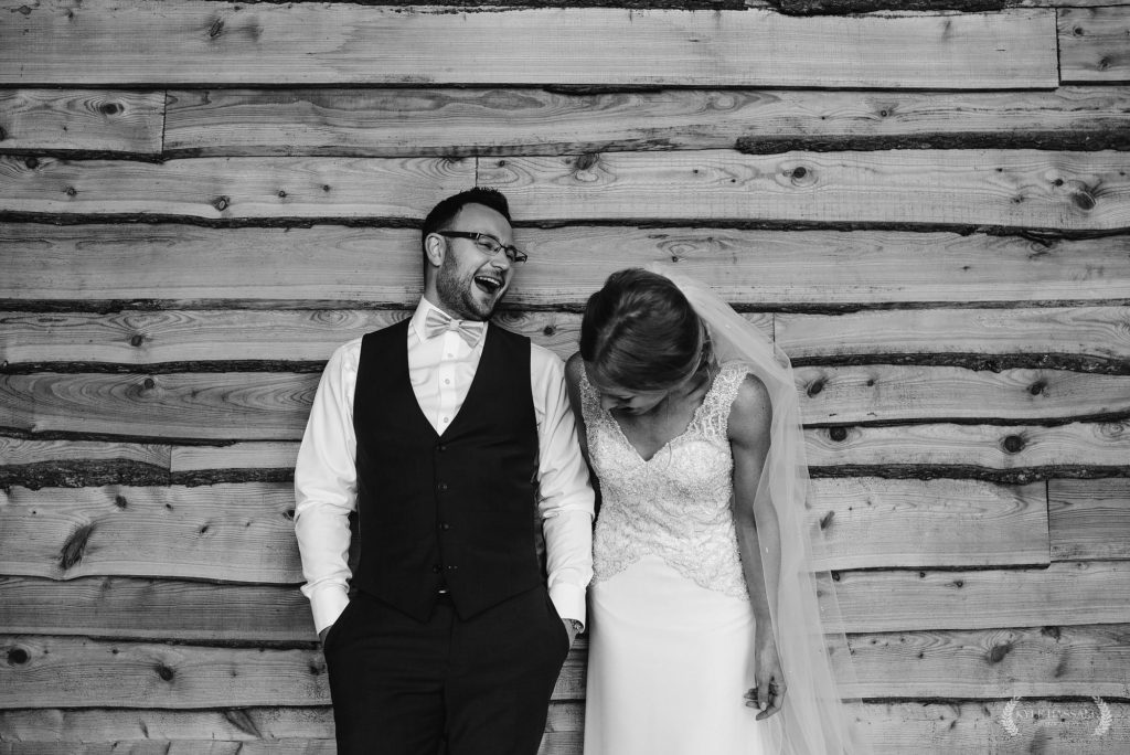 Bride and Groom laughing while leaning on a wooden wall