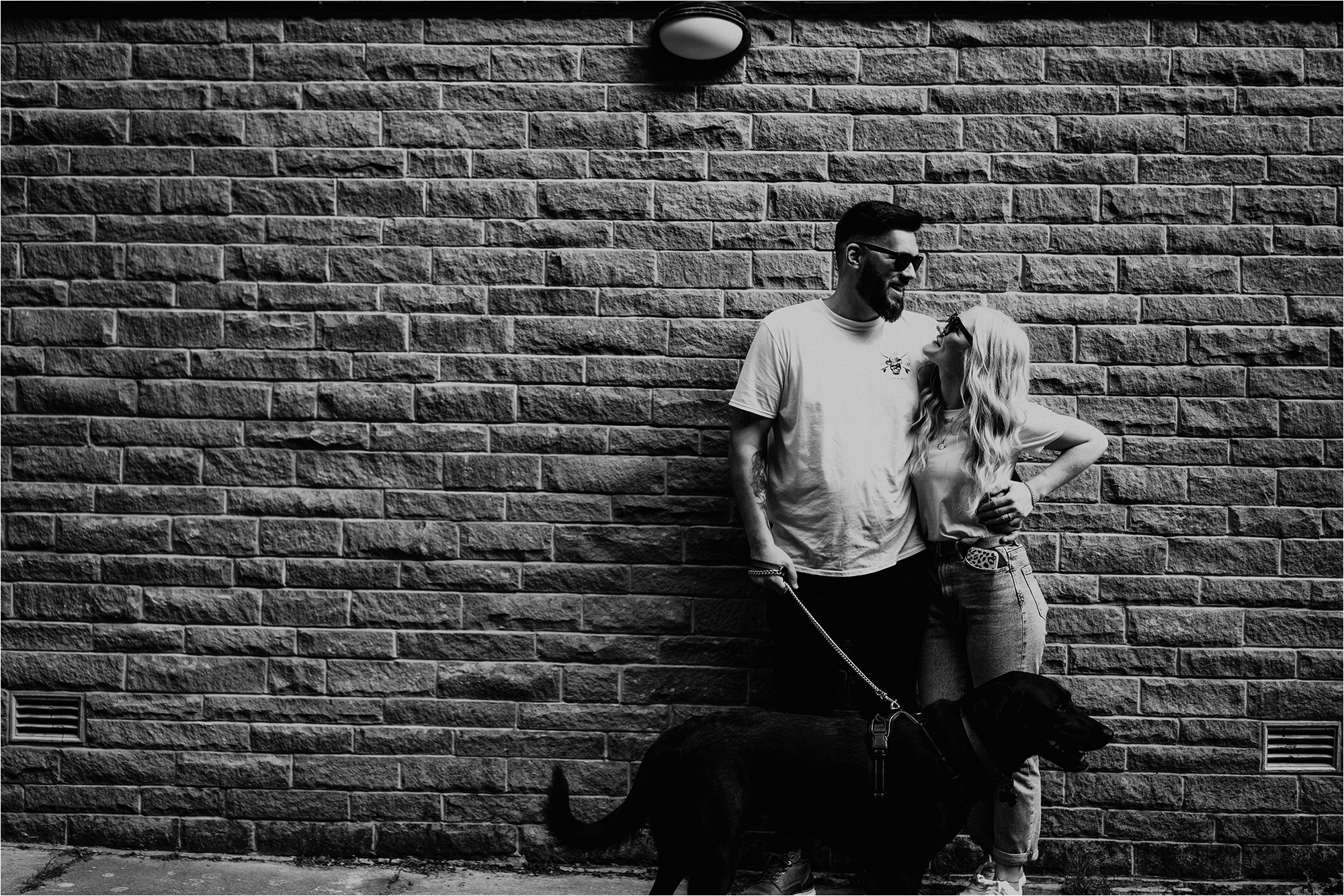 Black and white photo of couple leaning against wall with their dog